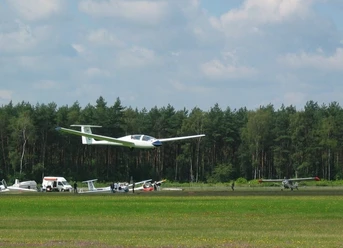 Segelflugplatz Segelflugzeug hebt vom grünen Flugplatz ab, im Hintergrund dichter Wald und ruhiger Himmel.
