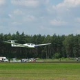 Segelflugplatz Segelflugzeug hebt vom grünen Flugplatz ab, im Hintergrund dichter Wald und ruhiger Himmel.