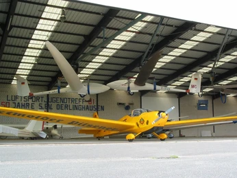 Segelflugplatz Gelbe Propellermaschine vor offener Halle, dahinter Segelflugzeuge an der Decke hängend.