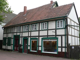 Haus Feldstraße Fachwerkhaus mit roten Ziegeln, grünen Fensterrahmen und zwei Schaufenstern, steht in einer ruhigen Straße.