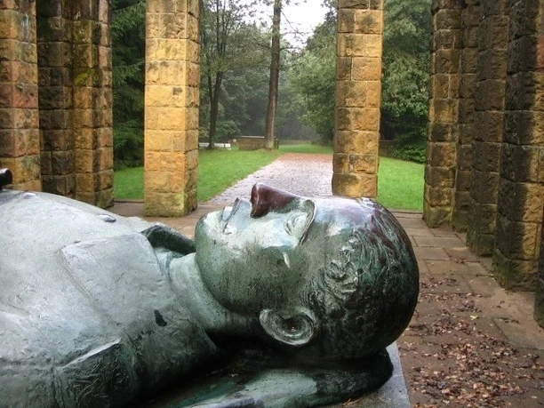 Ehrenmal Gedenkstätte mit liegender Steinskulptur, von Säulen umgeben, in einer bewaldeten Parklandschaft.