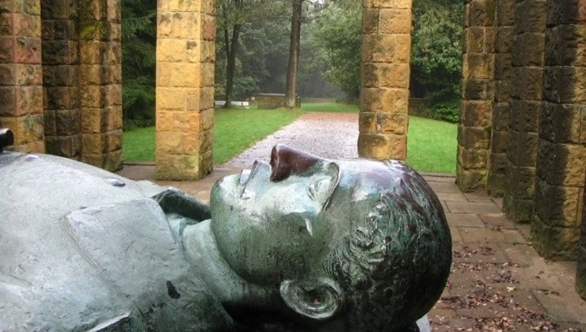 Ehrenmal Gedenkstätte mit liegender Steinskulptur, von Säulen umgeben, in einer bewaldeten Parklandschaft.