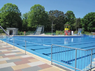 Schwimmbad mit einem großen rechteckigen Becken, umgeben von grünen Bäumen und einem Wasserrutsche.