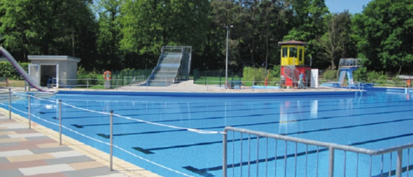 Schwimmbad mit einem großen rechteckigen Becken, umgeben von grünen Bäumen und einem Wasserrutsche.