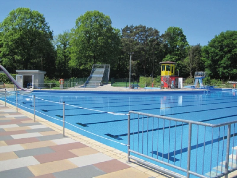 Schwimmbad mit einem großen rechteckigen Becken, umgeben von grünen Bäumen und einem Wasserrutsche.