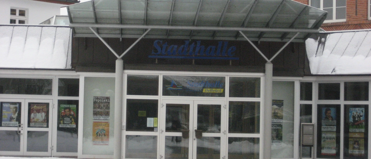 Eingangsbereich der Stadthalle Delbrück im Winter mit schneebedecktem Dach und Plakaten an der Glasfassade.