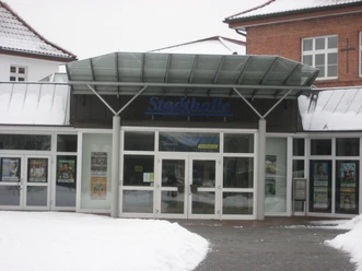 Stadthalle Delbrück Eingangsbereich der Stadthalle Delbrück im Winter mit schneebedecktem Dach und Plakaten an der Glasfassade.