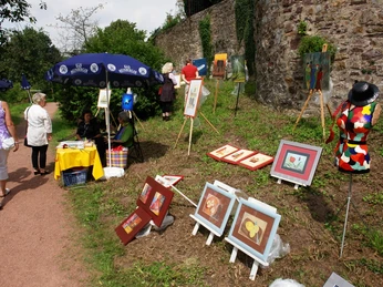 Kunstmauer am Weinberg, Outdoorgalerie am ersten Augustsonntag Bunte Kunstwerke auf Staffeleien an einer Steinmauer im Freien, betrachtet von Spaziergängern.
