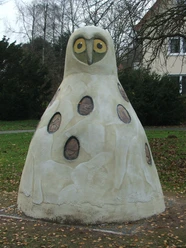 Horst von Hörste Große Beton-Eulenskulptur in einem Park, umgeben von Bäumen und einem Haus im Hintergrund.