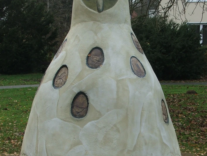 Horst von Hörste Große Beton-Eulenskulptur in einem Park, umgeben von Bäumen und einem Haus im Hintergrund.