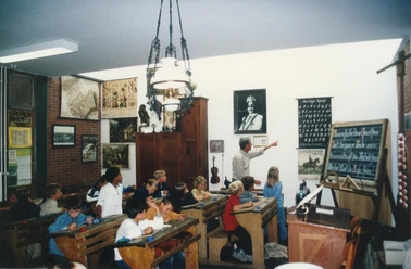 Ein Lehrer erklärt in einem historischen Klassenzimmer die Schulgeschichte, umgeben von Schautafeln.