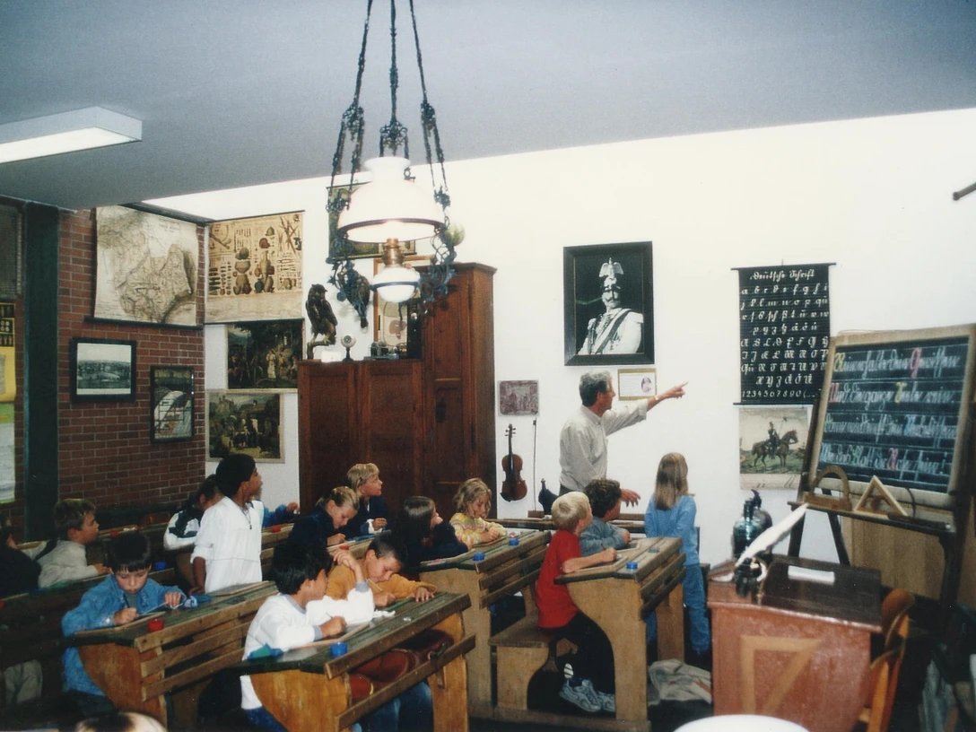 Ein Lehrer erklärt in einem historischen Klassenzimmer die Schulgeschichte, umgeben von Schautafeln.