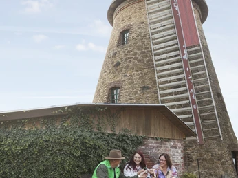 Rast an der Mühle Drei Personen entspannen bei einem Getränk an einem Holztisch vor einer historischen Windmühle.