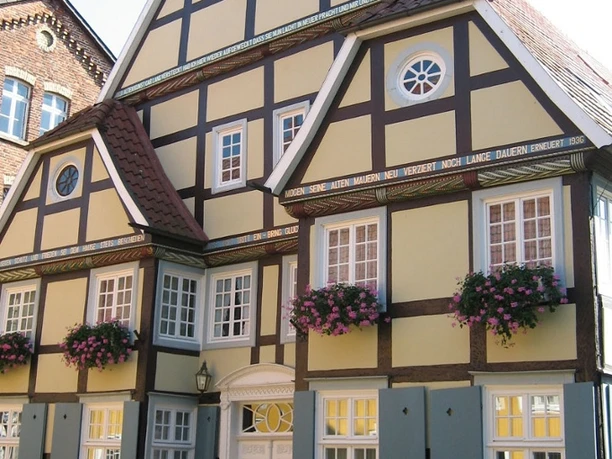 Fachwerkhaus mit weißen Fenstern, braunen Balken und rosa Blütenkästen, vor kleinem Gartenzaun.