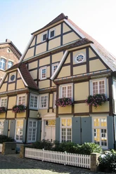 Rietberger Fachwerkhaus Fachwerkhaus mit weißen Fenstern, braunen Balken und rosa Blütenkästen, vor kleinem Gartenzaun.
