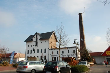 Ehemalige Kornbrennerei Meyer & WEZ Historische Backsteinbrennerei mit Schornstein, vor modernem Eingang platziert, flankiert von Autos.