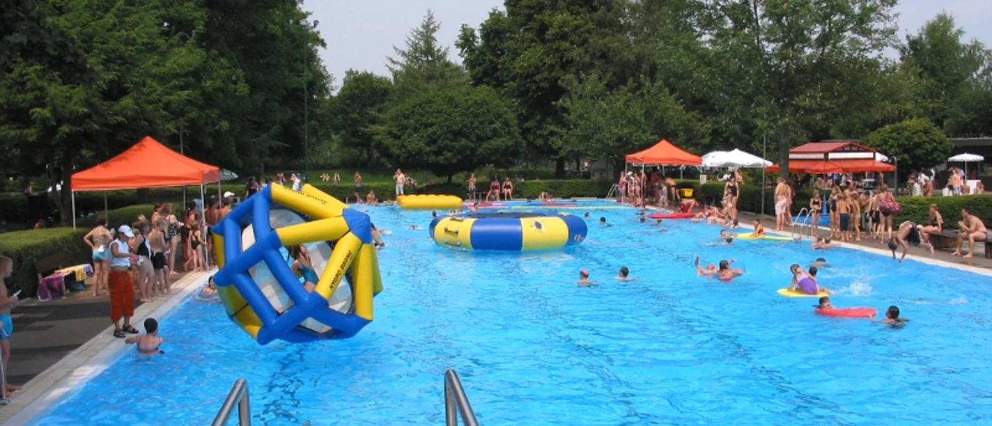 Ein lebhaftes Schwimmbad voller Menschen mit aufblasbaren Spielgeräten und grünen Bäumen im Hintergrund.