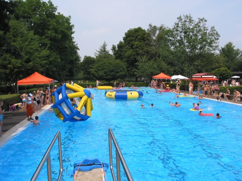 Ein lebhaftes Schwimmbad voller Menschen mit aufblasbaren Spielgeräten und grünen Bäumen im Hintergrund.