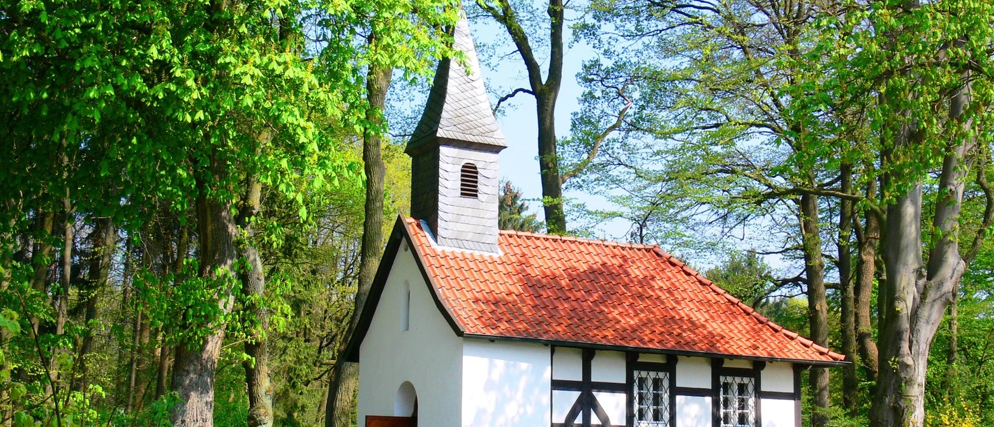 Weiße Kapelle mit rotem Ziegeldach, umgeben von grünen Bäumen und einem sonnigen Himmel.