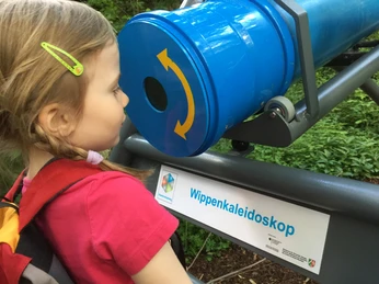 Ein Mädchen betrachtet gebannt ein großes blaues Kaleidoskop in einem grünen Outdoor-Bereich.