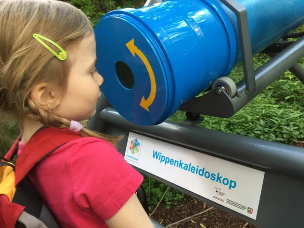 Nach dem Herabbewegen schauen Sie in das Kaleidoskop Ein Mädchen betrachtet gebannt ein großes blaues Kaleidoskop in einem grünen Outdoor-Bereich.
