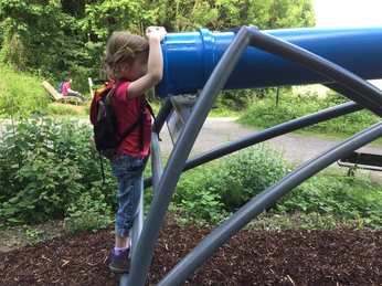 Am Sphäriskop laden eine Liegebank und eine Sitzbank zur Rast ein Ein Mädchen mit Rucksack blickt durch ein blaues Sphäriskop; daneben eine Sitzgelegenheit im Grünen.