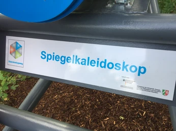 Das Spiegelkaleidoskop ist Teil des Kaleidoskopwegs Schildtext "Spiegelkaleidoskop" am Kaleidoskopweg, umgeben von grünem Laub und Holzboden.