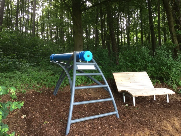 Zeitlupenkaleidoskop auf Stahlgestell neben einer hölzernen Liegebank in einem bewaldeten Bereich.