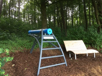 Das Zeitlupenkaleidoskop steht direkt neben einer Liegebank Zeitlupenkaleidoskop auf Stahlgestell neben einer hölzernen Liegebank in einem bewaldeten Bereich.