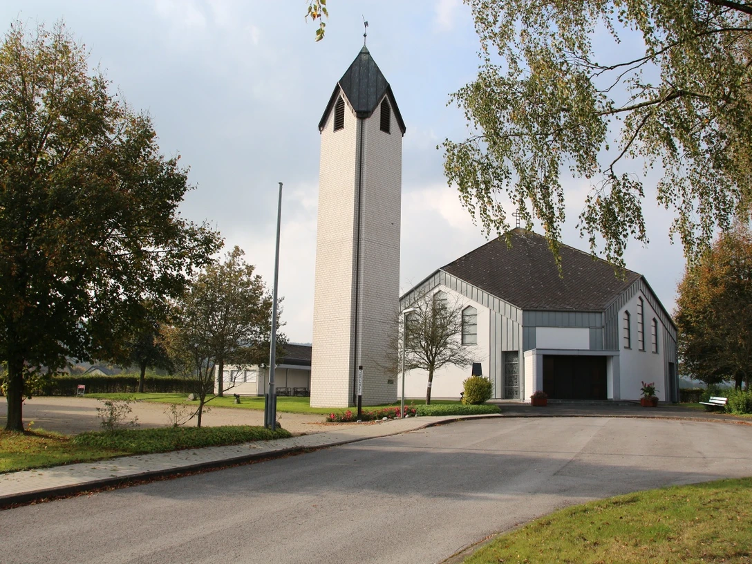 Die Johannes Nepomuk Kirche mit weiß-grauem Turm und Vorplatz, umgeben von Bäumen und ruhiger Umgebung.