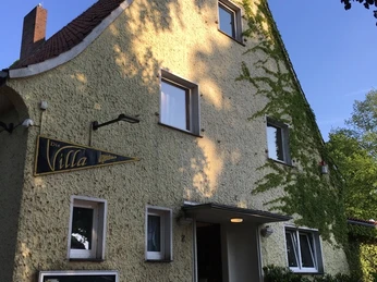 Jugendhaus "Die Villa" von vorne Gelbes, mit Efeu bewachsenes Gebäude unter blauem Himmel. Fahrräder parken vor dem Eingang.