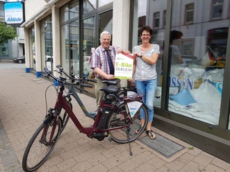 Ein Paar steht lächelnd neben einem E-Bike vor einem Verleih mit Ausschilderung, tagsüber.