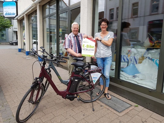 movelo E-Bike Verleih Betten- Paradies & Pension Schübeler Ein Paar steht lächelnd neben einem E-Bike vor einem Verleih mit Ausschilderung, tagsüber.