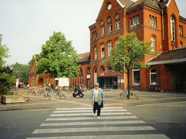 Roter Backsteinbahnhof mit Bäumen, Fahrradständern und einer Person auf einem Zebrastreifen davor.