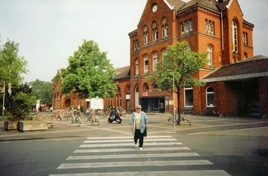 Bahnhof Detmold Roter Backsteinbahnhof mit Bäumen, Fahrradständern und einer Person auf einem Zebrastreifen davor.