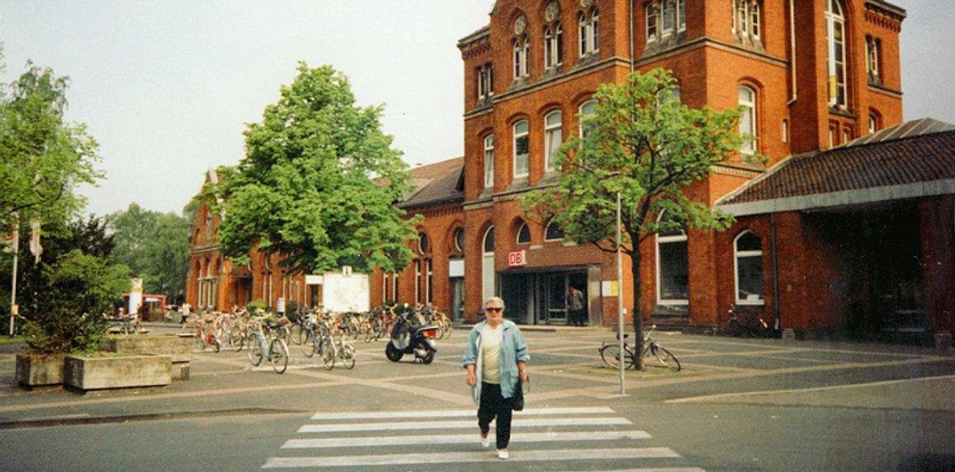 Bahnhof Detmold Roter Backsteinbahnhof mit Bäumen, Fahrradständern und einer Person auf einem Zebrastreifen davor.