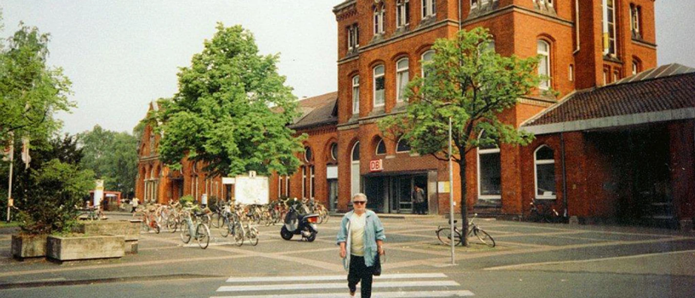 Bahnhof Detmold Roter Backsteinbahnhof mit Bäumen, Fahrradständern und einer Person auf einem Zebrastreifen davor.
