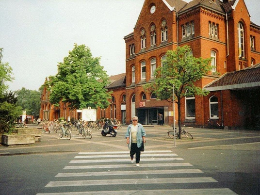 Bahnhof Detmold Roter Backsteinbahnhof mit Bäumen, Fahrradständern und einer Person auf einem Zebrastreifen davor.