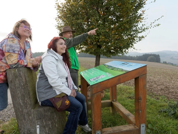 Panoramatafel mit Blick auf Talle Drei Personen betrachten eine Panoramatafel auf einem Feldweg mit Aussicht auf eine hügelige Landschaft.