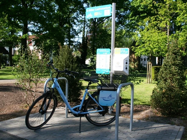 Fahrrad steht an einer Ladestation für E-Bikes im sonnigen Park, umgeben von grünen Bäumen.