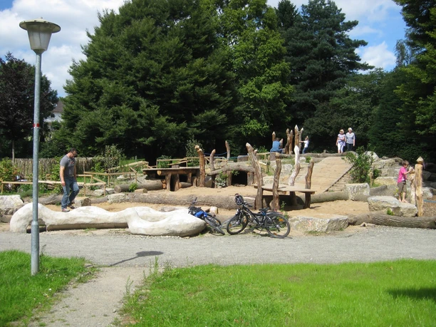 Spielplatz mit natürlichen Elementen wie Holzskulpturen und Sand, umgeben von Bäumen und Besuchern.