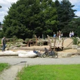 image Spielplatz mit natürlichen Elementen wie Holzskulpturen und Sand, umgeben von Bäumen und Besuchern.