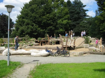 image Spielplatz mit natürlichen Elementen wie Holzskulpturen und Sand, umgeben von Bäumen und Besuchern.