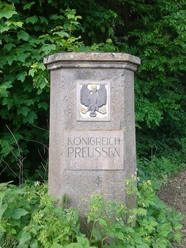 Steinerner Hoheitsstein in Harzberg mit preußischem Adler, umgeben von dichter Vegetation.