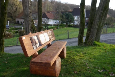 "Adel und Hochmut" Holzbank mit geschnitzter Rückenlehne, daneben ein Weg und Fachwerkhäuser im Hintergrund.