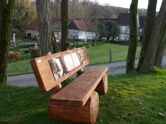 "Adel und Hochmut" Holzbank mit geschnitzter Rückenlehne, daneben ein Weg und Fachwerkhäuser im Hintergrund.
