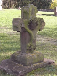 Steinernes Kreuz mit Jesusrelief in grüner Wiese, von Moos bewachsen, vor einem bewaldeten Hintergrund.