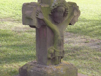 Scharfrichter Kreuz Steinernes Kreuz mit Jesusrelief in grüner Wiese, von Moos bewachsen, vor einem bewaldeten Hintergrund.