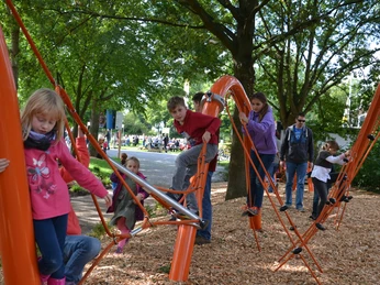 Kinder klettern auf orangefarbenem Spielgerüst im Park, umgeben von grünen Bäumen und hellem Sonnenschein.