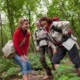 Eine Gruppe von drei Wanderern lacht auf einem Waldweg, umgeben von grünen Bäumen und Pflanzen.